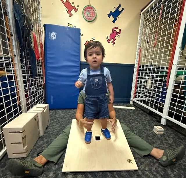 A young child practising standing on a board with a therapist supporting from behind
