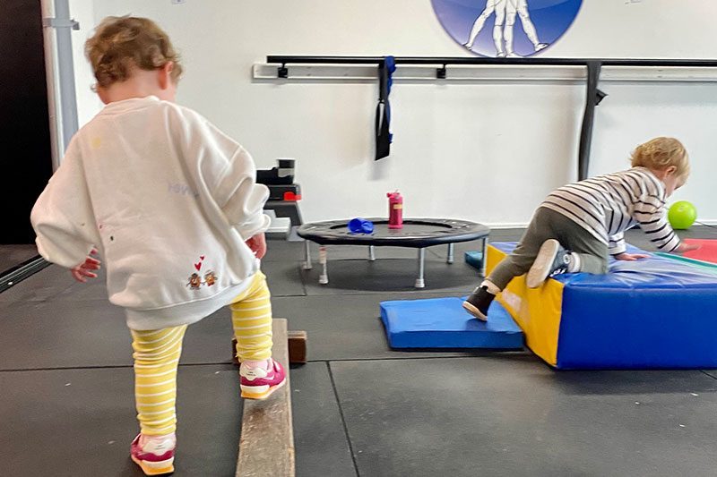 Two toddlers moving over beams and soft blocks in a therapy space